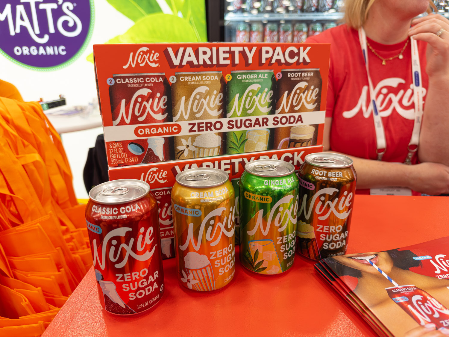 A Nixie zero sugar soda variety pack and loose cans on a red display with a person in a Nixie shirt behind