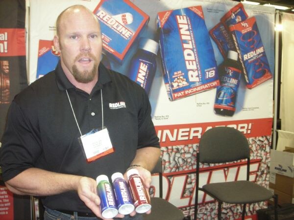 A bald man with a goatee in a black Redline shirt holds three small Redline bottles in front of a backdrop of Redline product graphics, with a chair beside him.