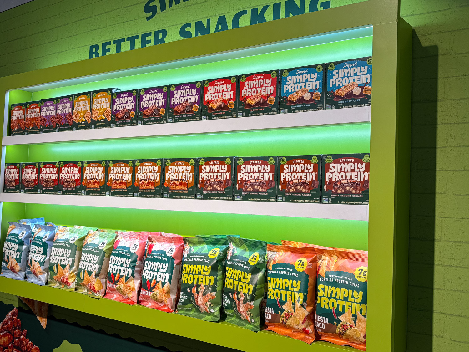 A green lit shelf wall fully stocked with Simply Protein snack bags