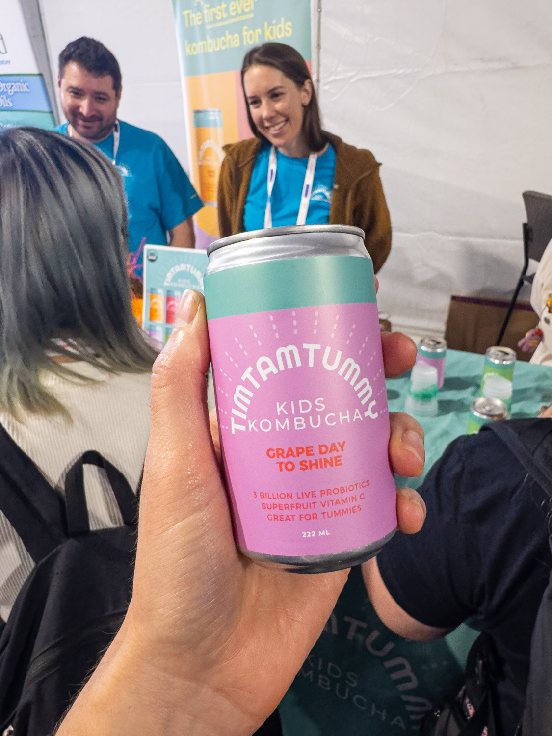 A hand holding a pink TimTamTummy kids kombucha can in front of two smiling people at a booth