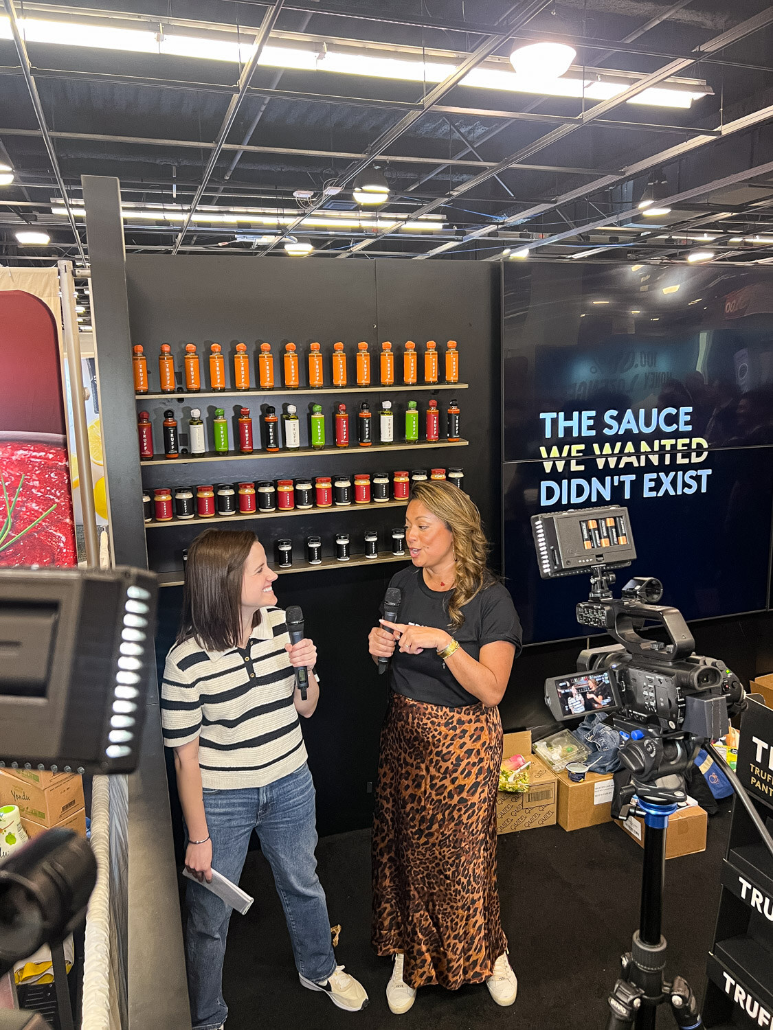Two women talking at a booth with shelves of hot sauce bottles while a camera records