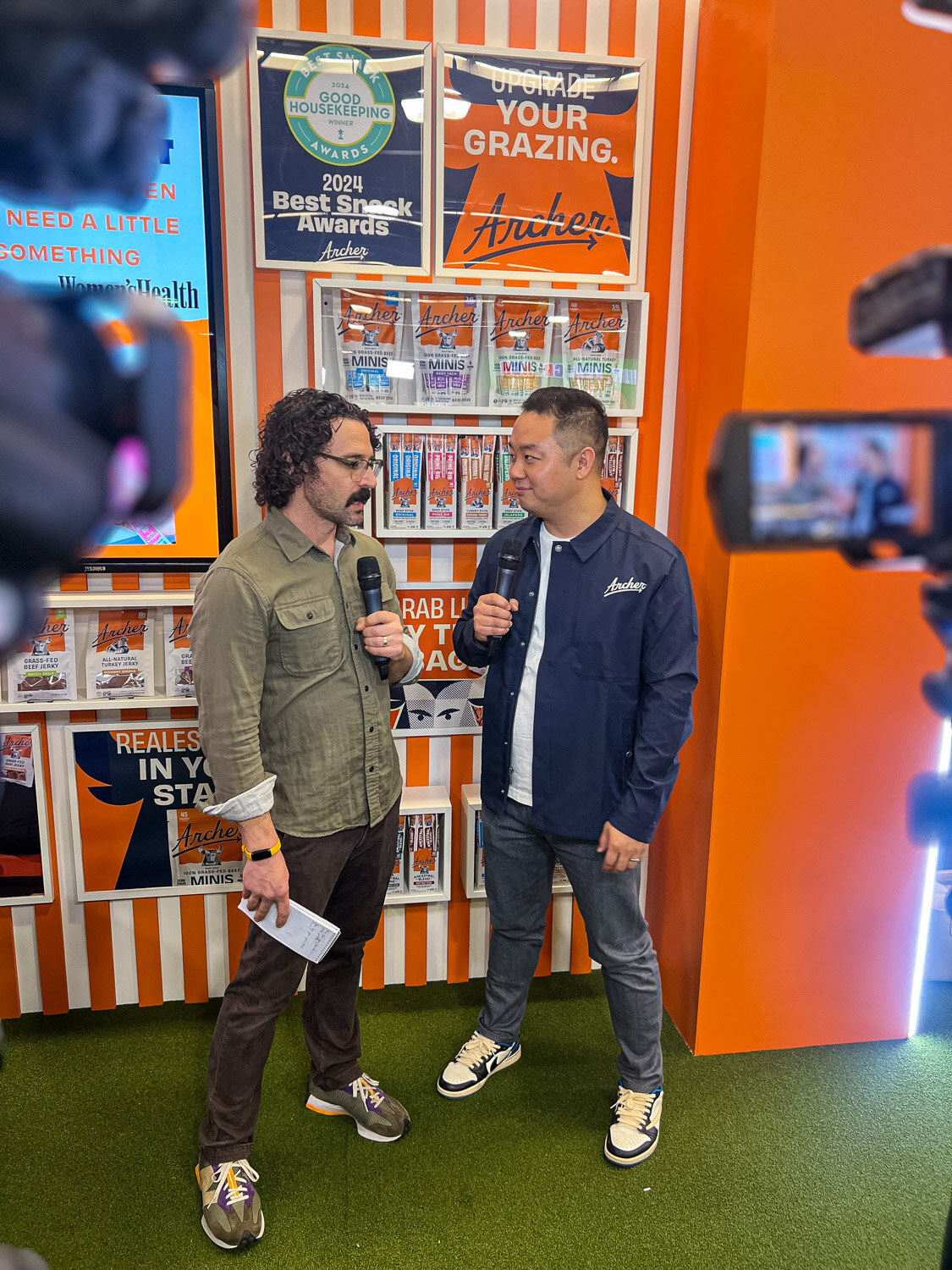 Two men in conversation holding a microphone in front of an orange and white booth while being filmed