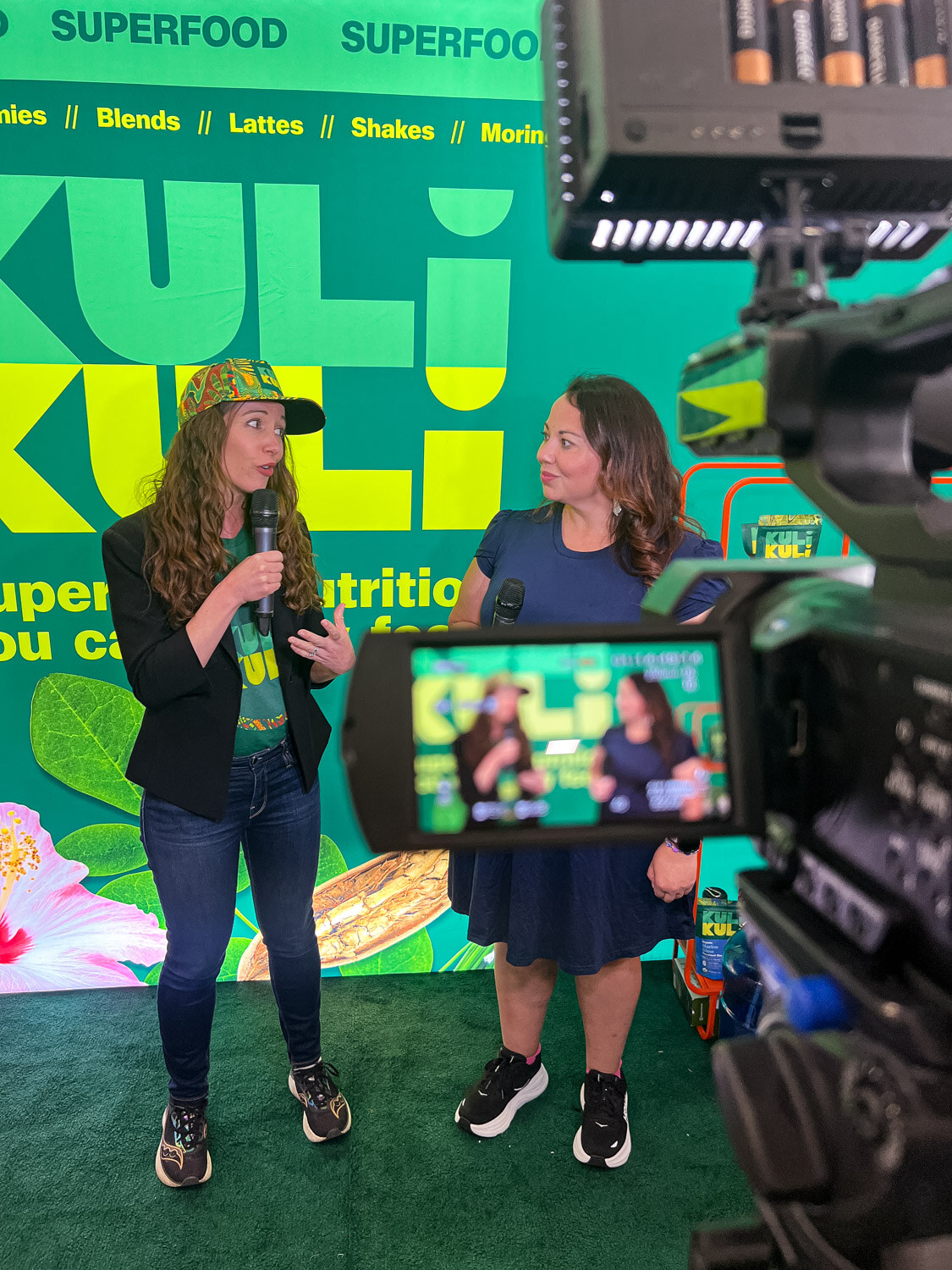 Two women talking in front of a green Kuli Kuli backdrop with a camera monitor showing the shot