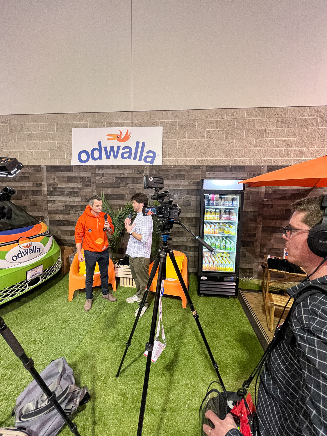 An interview being filmed on artificial grass in front of an Odwalla sign with cameras on tripods