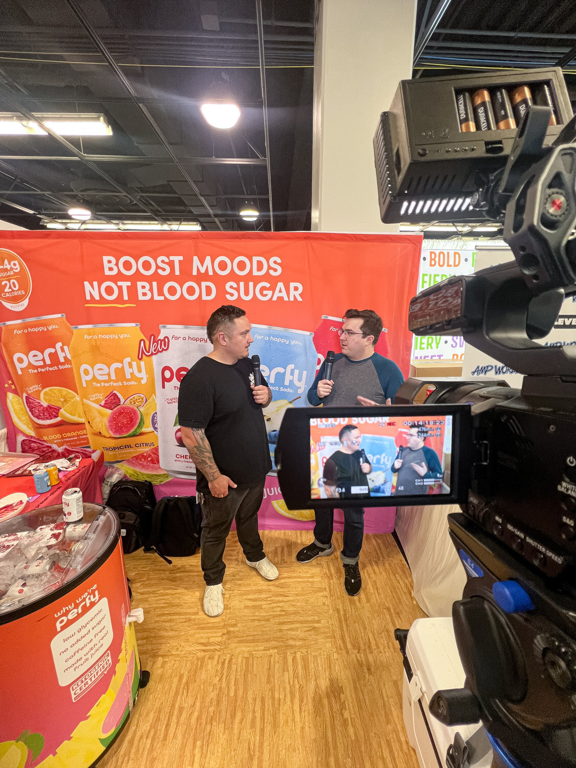 Two men talking on a small set in front of an orange Perky Boost Moods sign while a camera monitor shows the shot