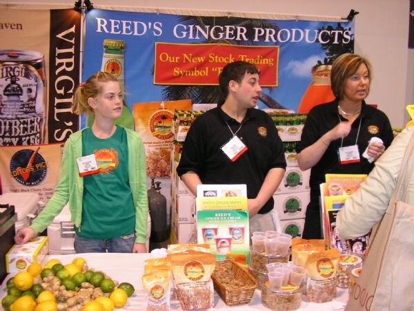 Three booth staff in black and green shirts stand behind a table under a Reed's Ginger Products banner, with a Virgil's banner at left and bags of ginger candy and fruit on the table.