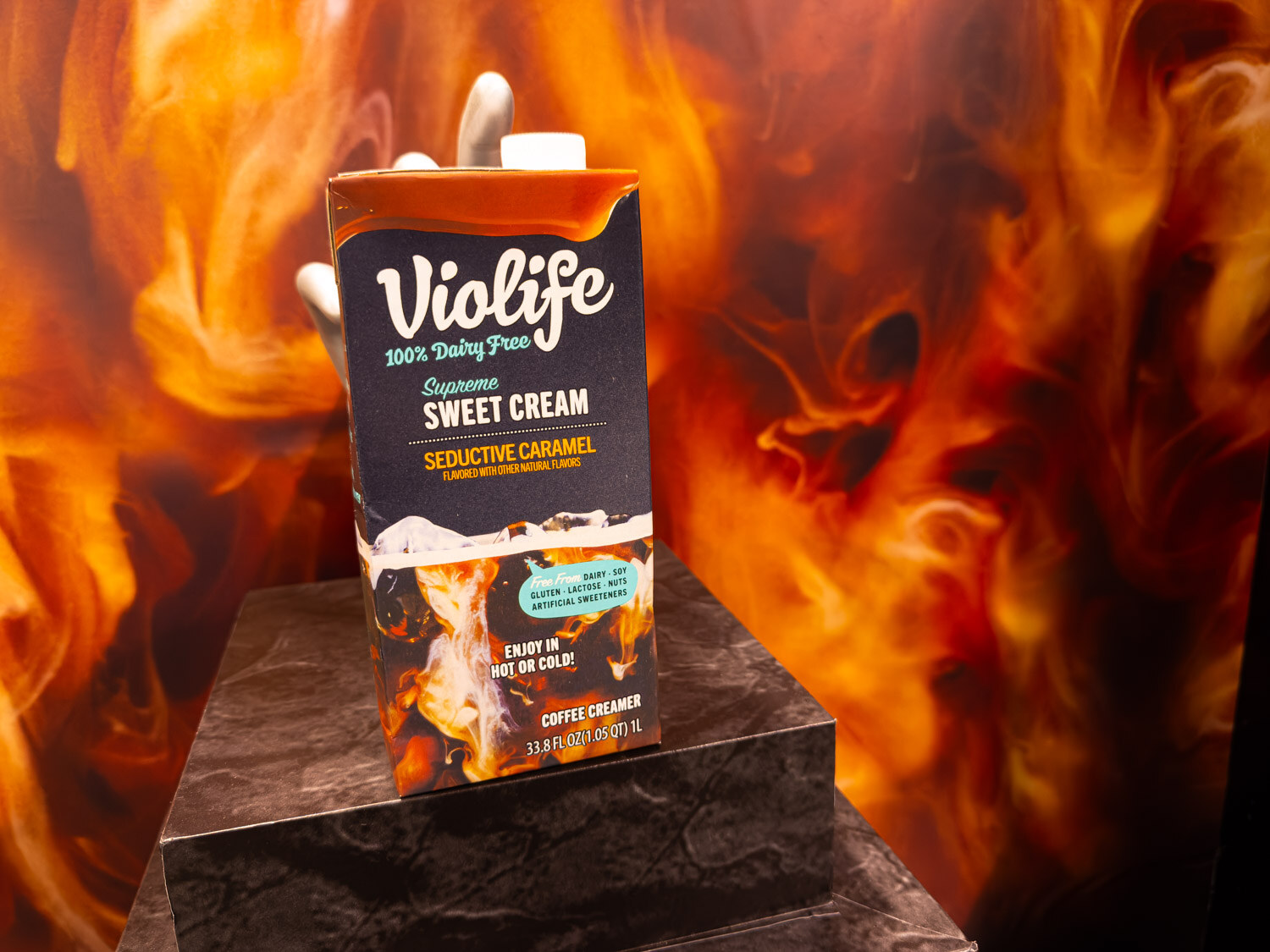 A Violife Supreme Sweet Cream coffee creamer carton on a black pedestal in front of a flame backdrop