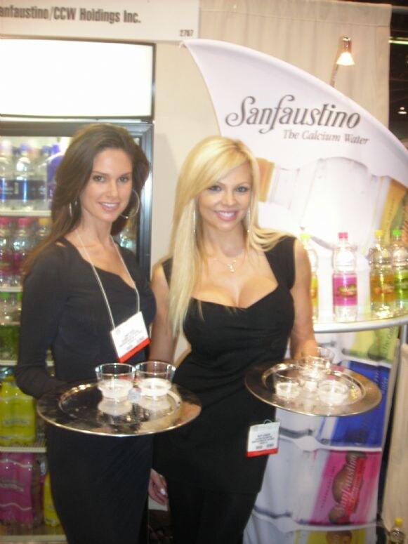 Two women in black dresses hold silver trays of small cups of water in front of a Sanfaustino The Calcium Water display. The booth sign reads Sanfaustino/CCW Holdings Inc.