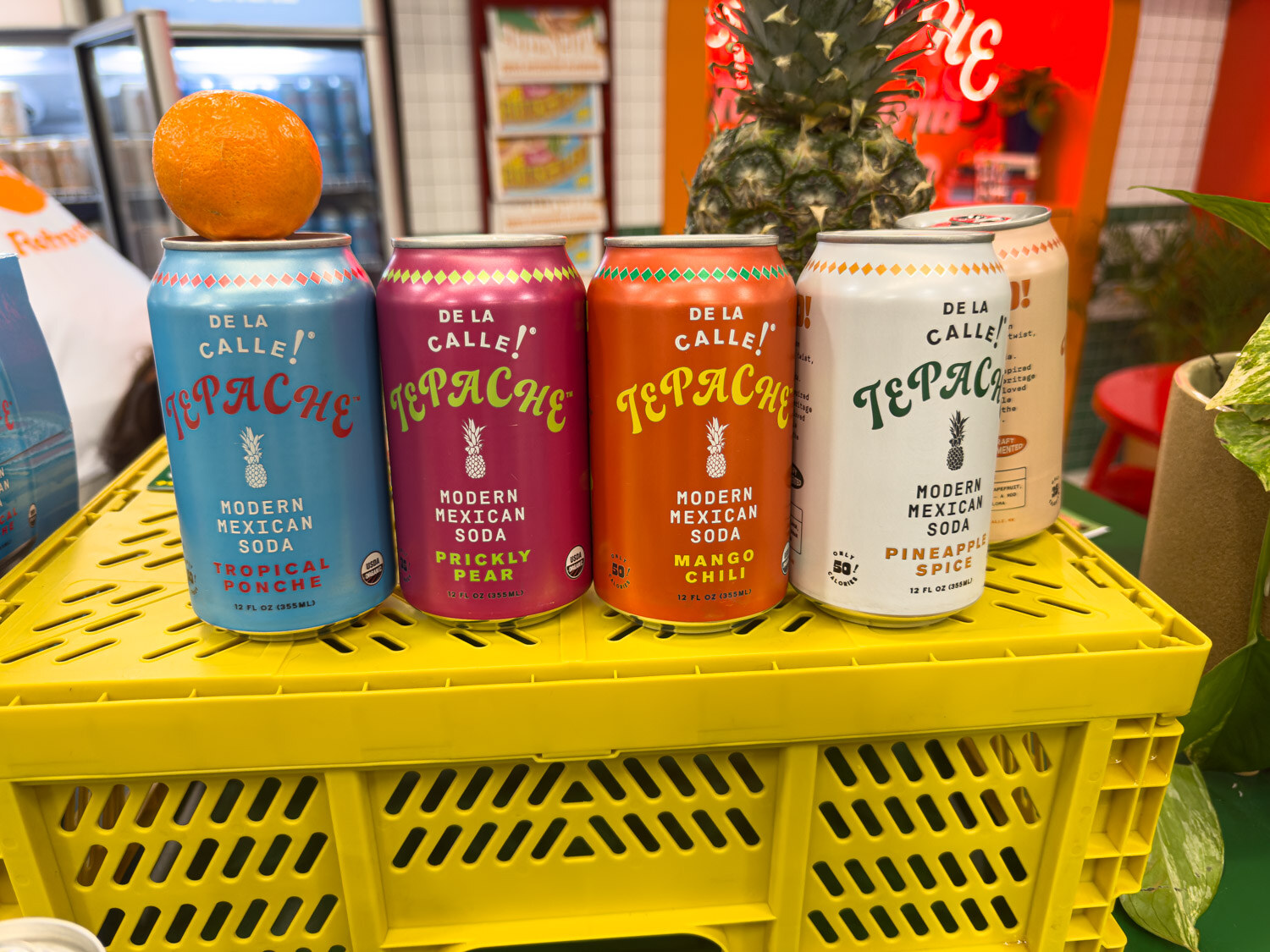 Four De La Calle Tepache cans on a yellow crate with an orange and a pineapple behind them