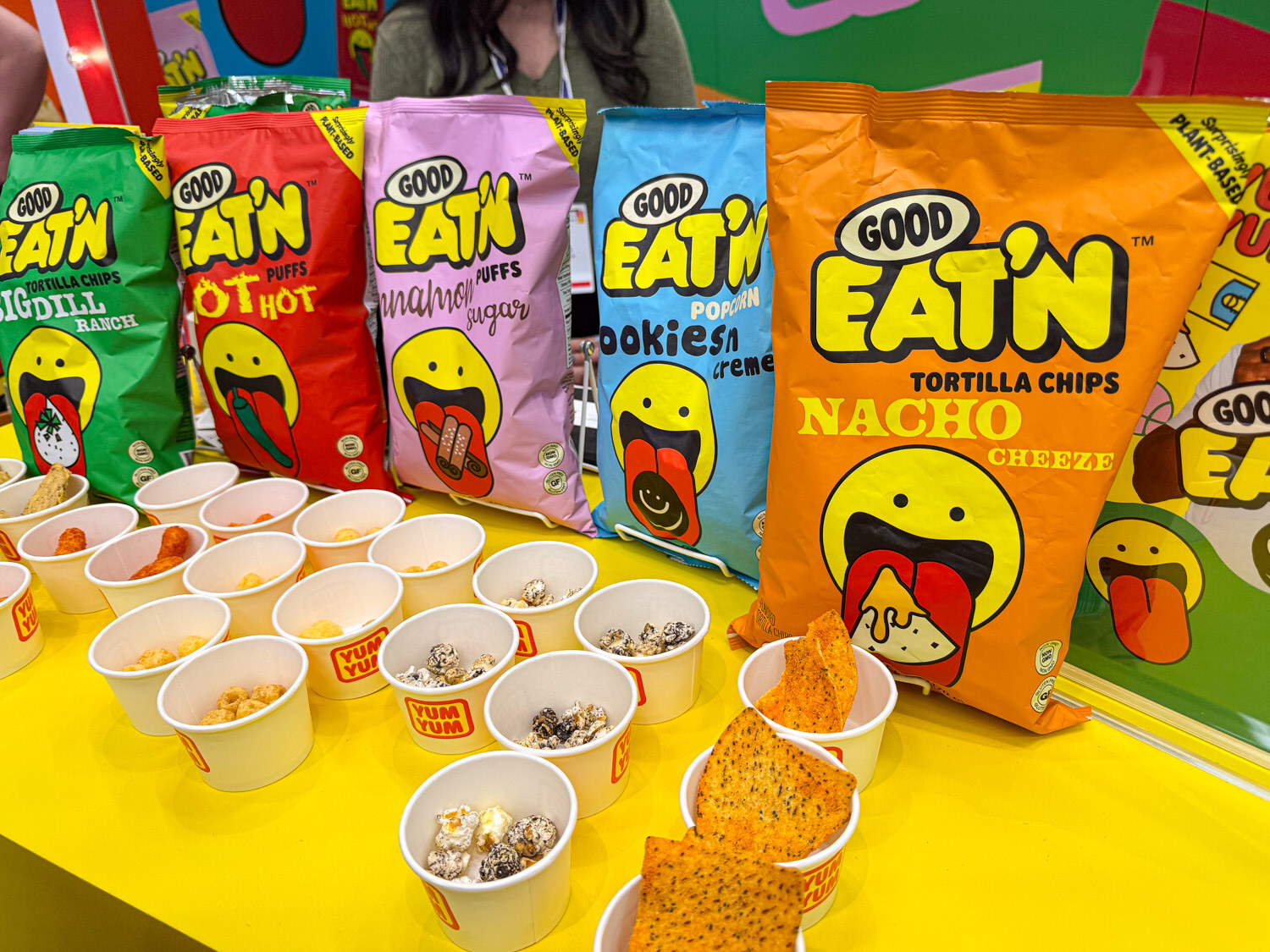 Good Eat'n tortilla chip and puff bags with sample cups on a yellow table