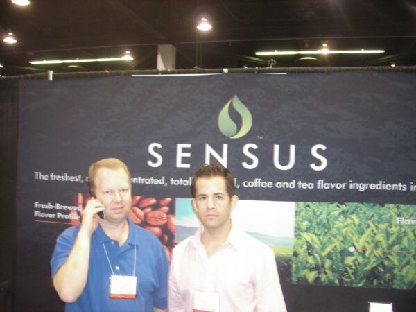 Two men stand in front of a dark Sensus banner showing coffee beans and tea leaves. The man at left, in a blue polo, holds a phone to his ear. The man at right wears a pink shirt.