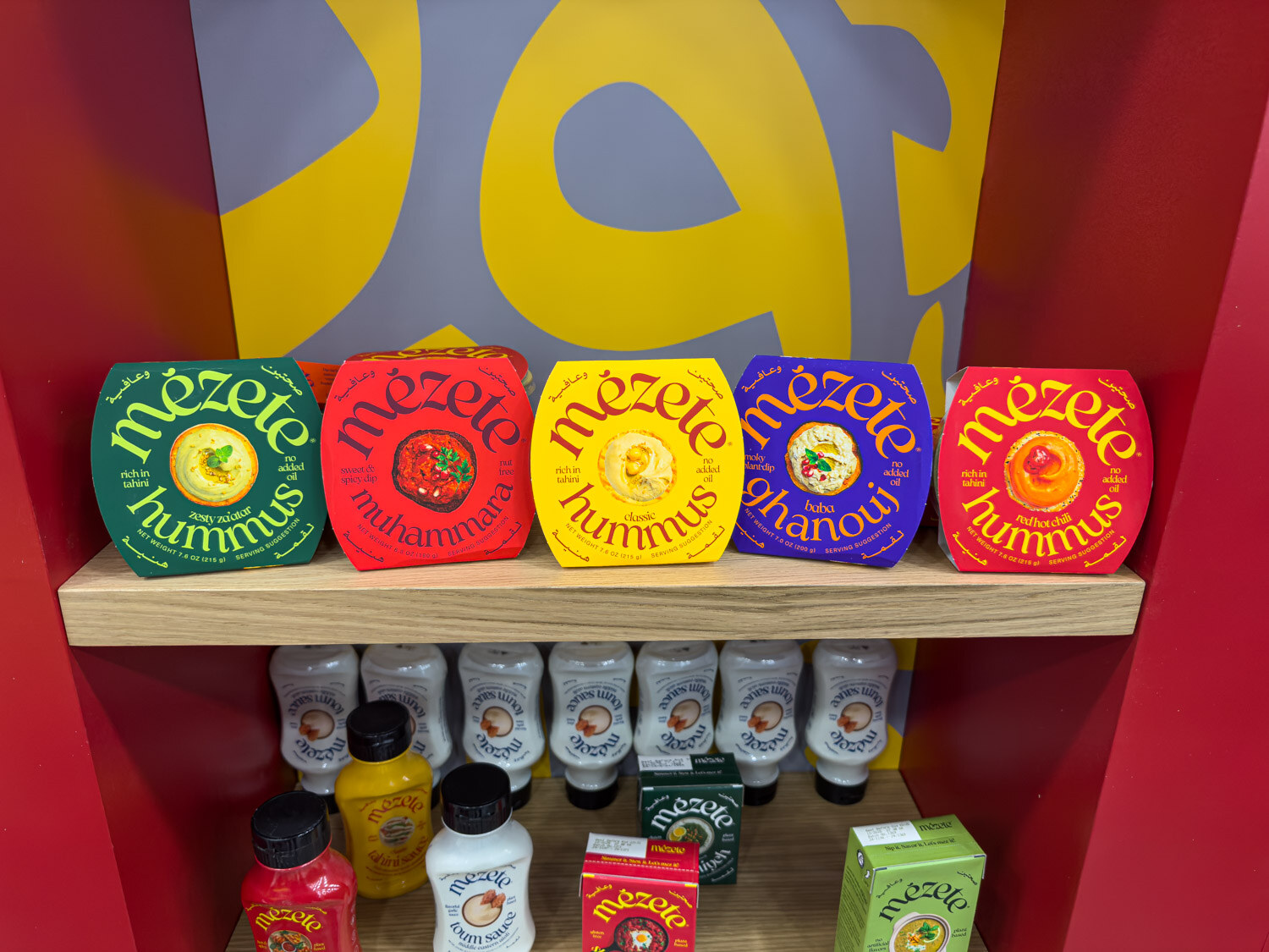 Mezete hummus tubs and squeeze sauces on a wooden shelf at a bright red booth