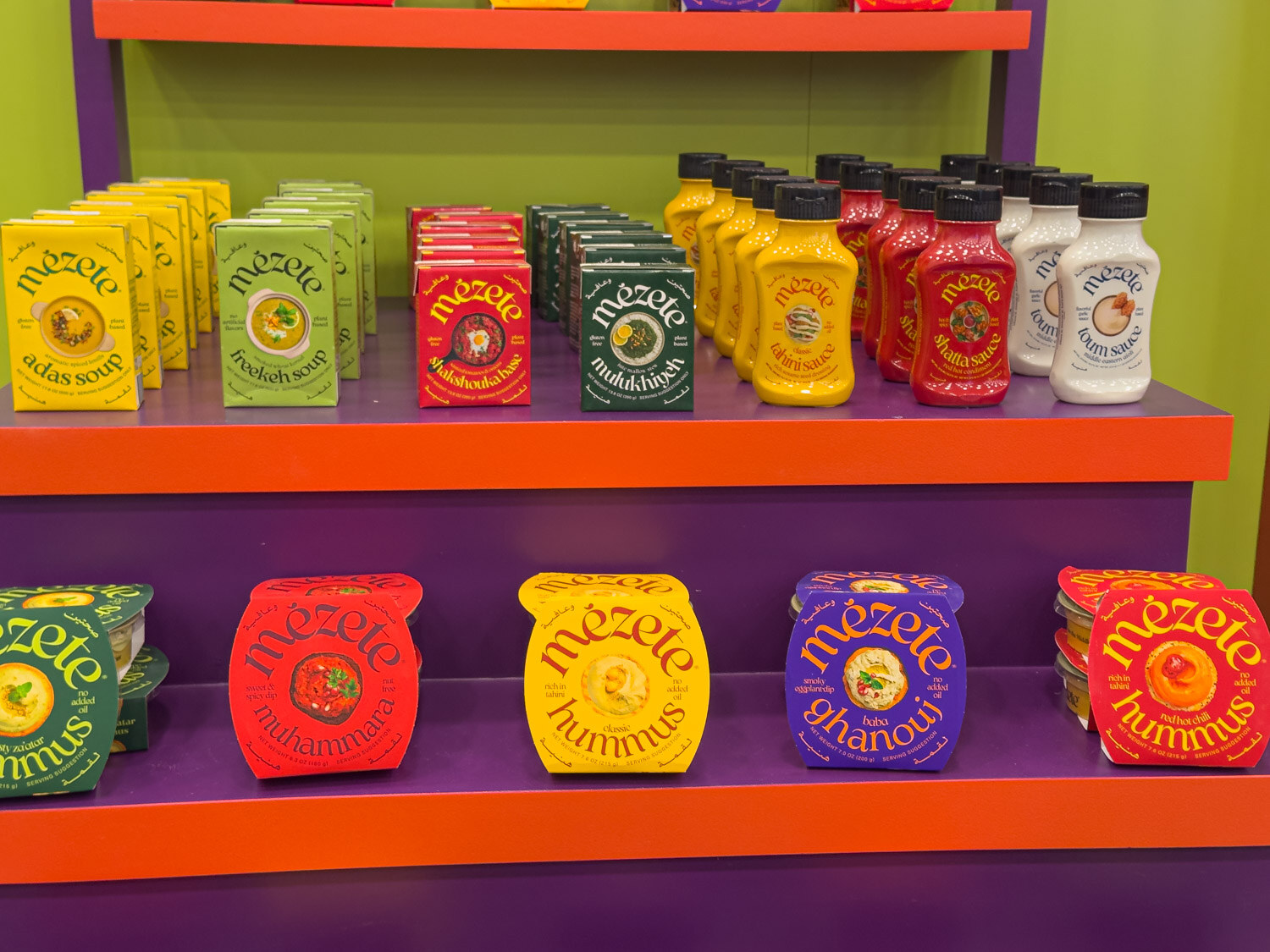 Mezete squeeze sauces, spreads and hummus tubs on tiered orange and purple shelves