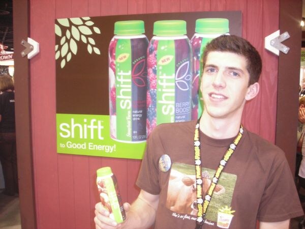 A young man in a brown T-shirt and lanyard holds a small green-capped bottle in front of a Shift to Good Energy sign showing bottles of berry drinks, on a red wooden wall.