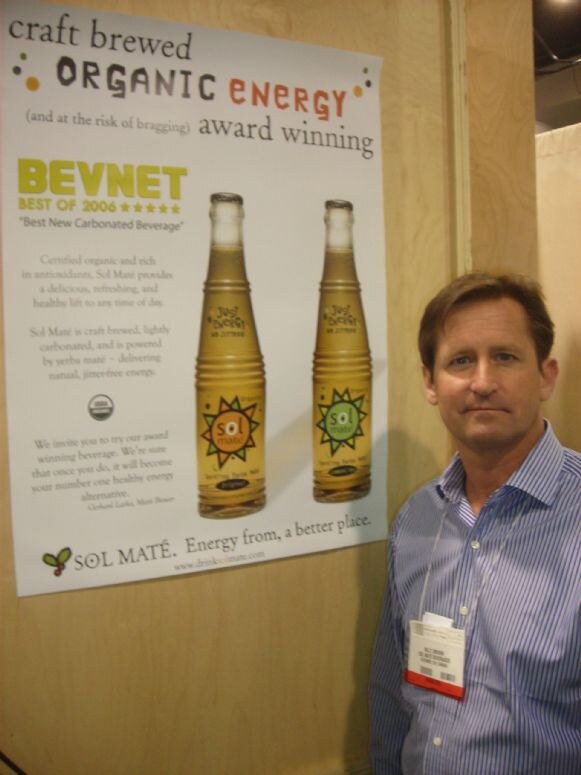 A man in a blue striped shirt with a show badge stands beside a Sol Maté poster. The poster has two bottles and text reading craft brewed organic energy and BevNET Best of 2006.