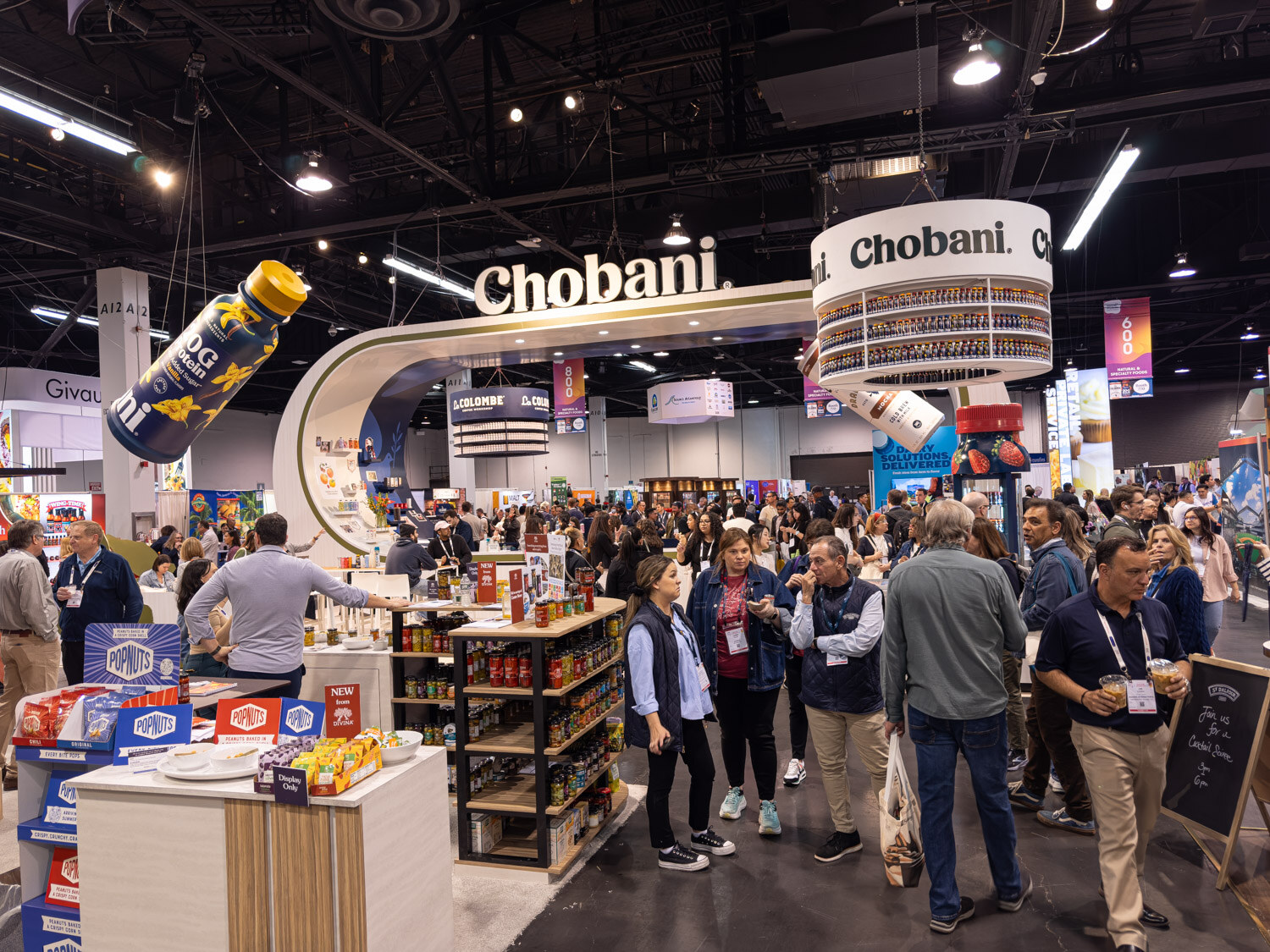 A Chobani booth with a curved sign and an oversized bottle hanging above a crowd of attendees