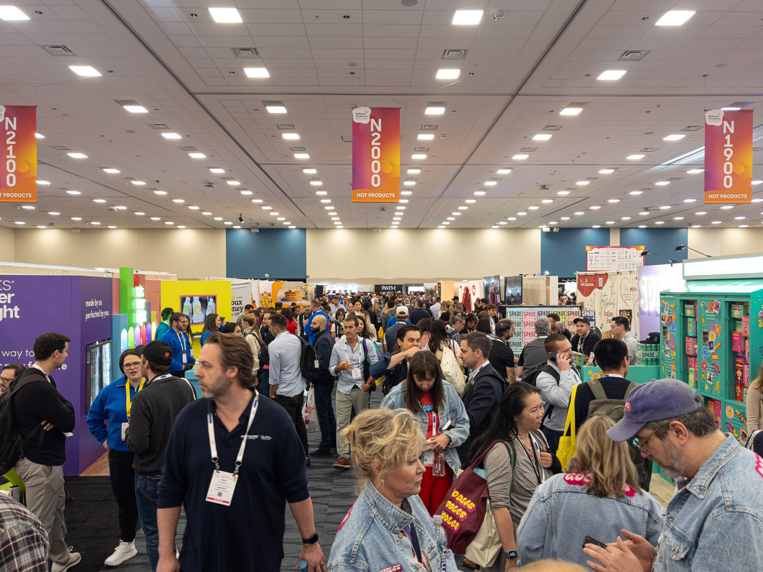 A busy exhibit hall with attendees walking between colorful booths under overhead aisle banners