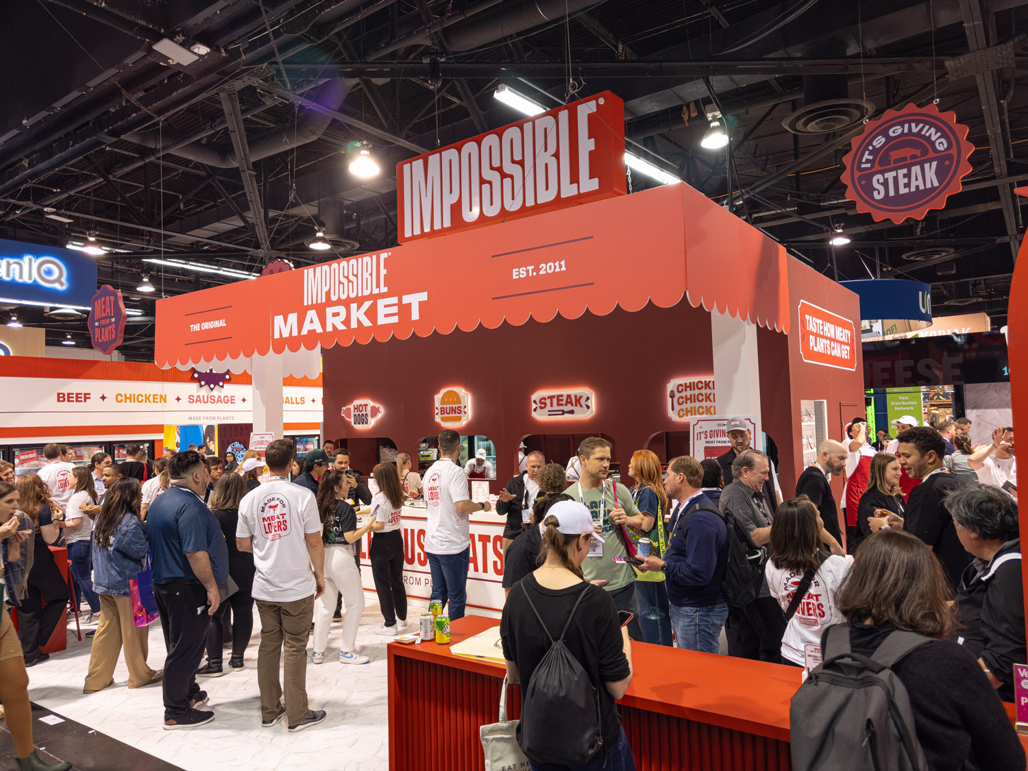 A large red Impossible Market booth surrounded by attendees on the exhibit floor