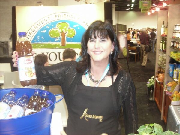 A smiling woman with dark hair in a black Source Atlantique Inc apron holds up a bottle of iced tea. A blue tub of bottled drinks sits in front of her.