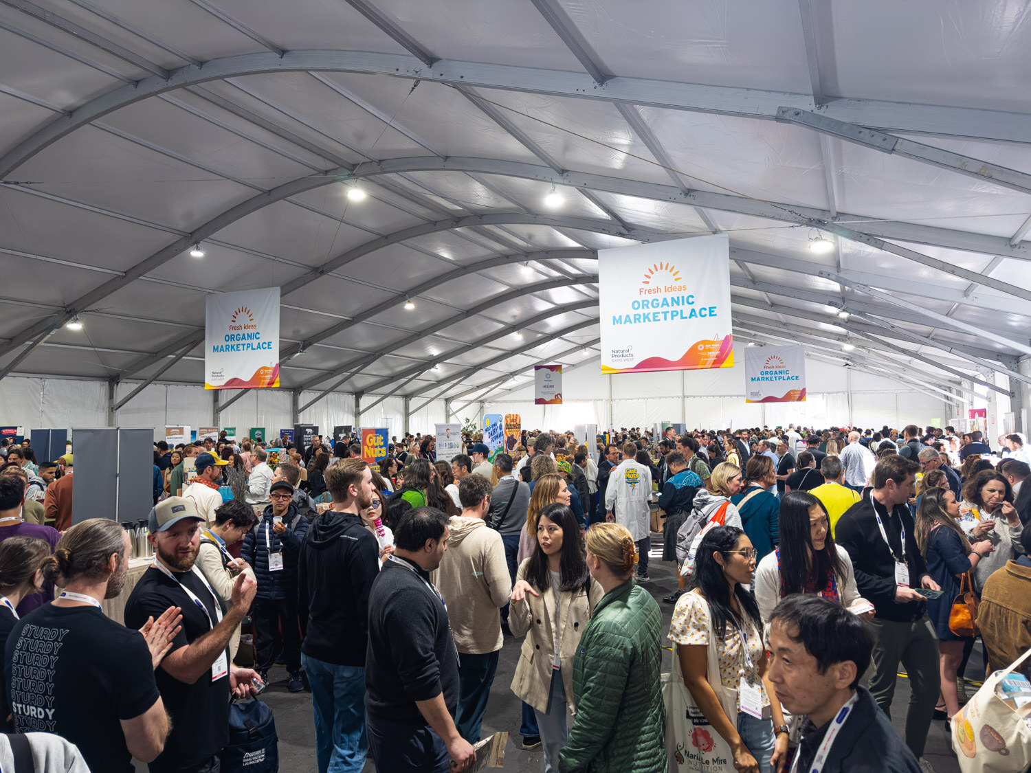 A crowded exhibition tent with an Organic Marketplace banner hanging from a curved white ceiling
