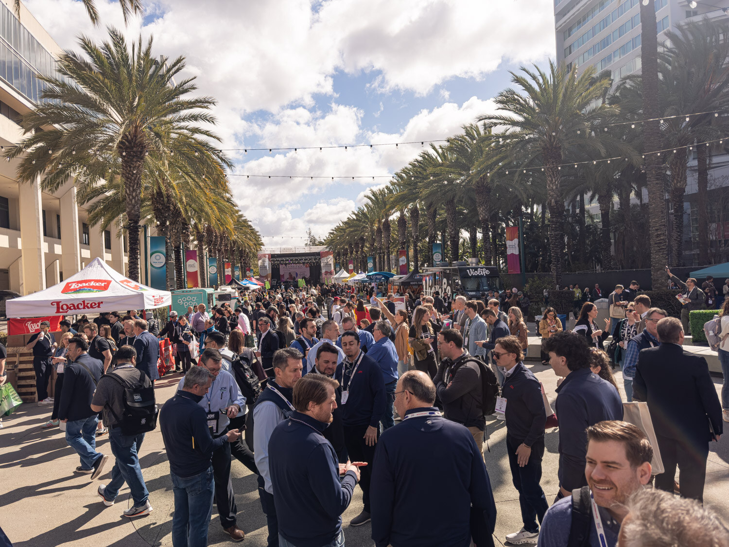 A large crowd of attendees on the palm-lined plaza outside the convention center under a blue sky