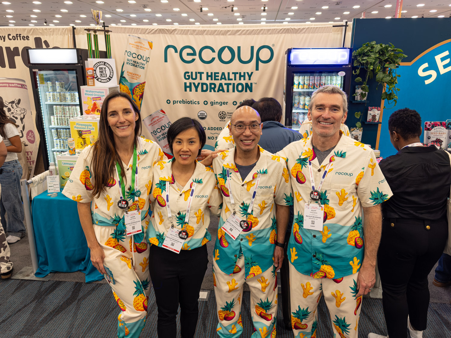 Four people in matching pineapple-print outfits smiling in front of a Recoup Gut Healthy Hydration booth