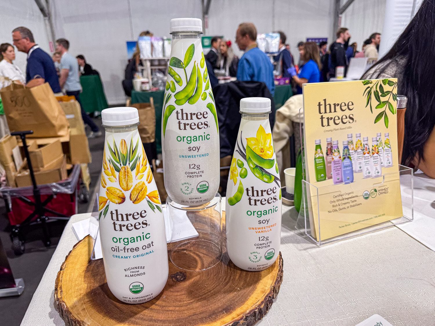 Three glass bottles of Three Trees organic soy and oat milks on a wooden slice with a product card behind them