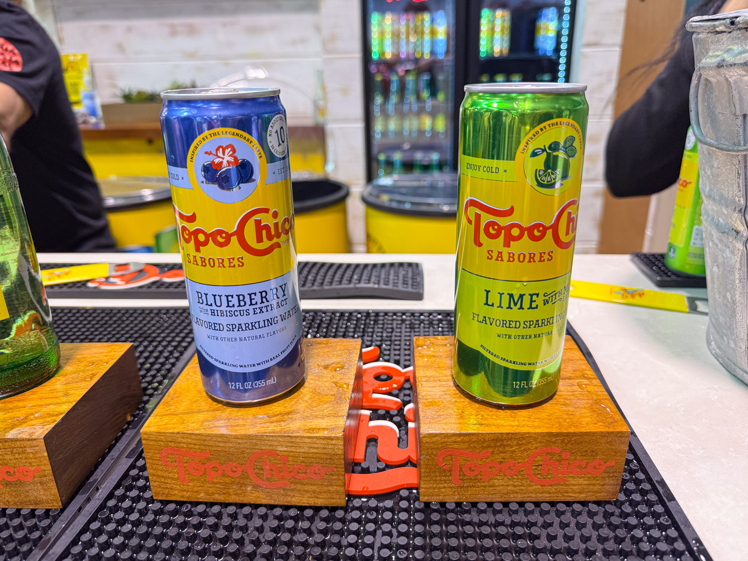 A blueberry and a lime Topo Chico Sabores can on wooden risers over a black bar mat
