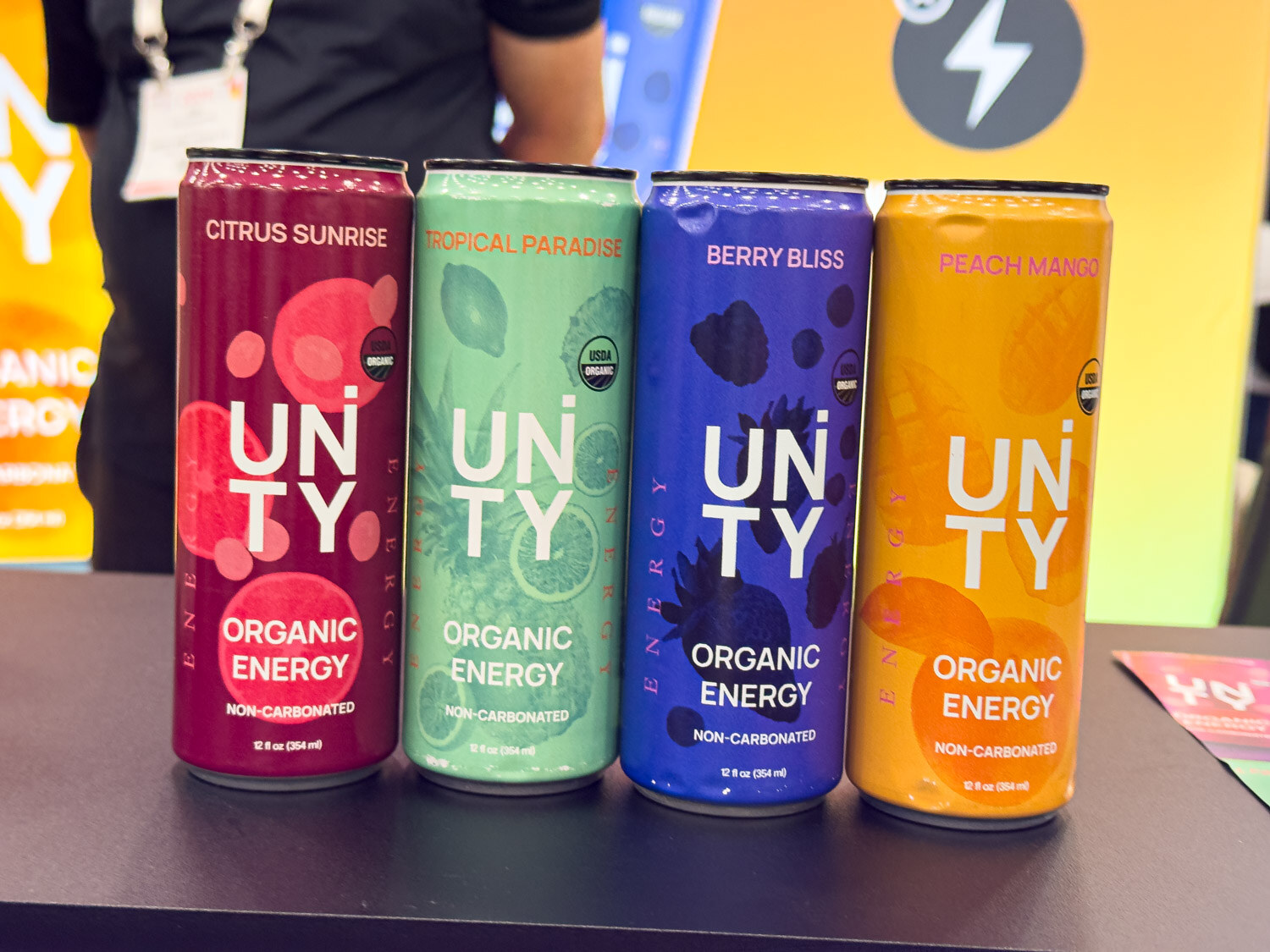 Four Unity organic energy drink cans in citrus sunrise, tropical paradise, berry bliss and peach mango
