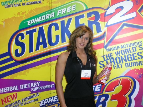 A woman with long brown hair, in a black sleeveless top with a show badge, holds a bottle in front of a yellow Stacker 2 Ephedra Free backdrop.
