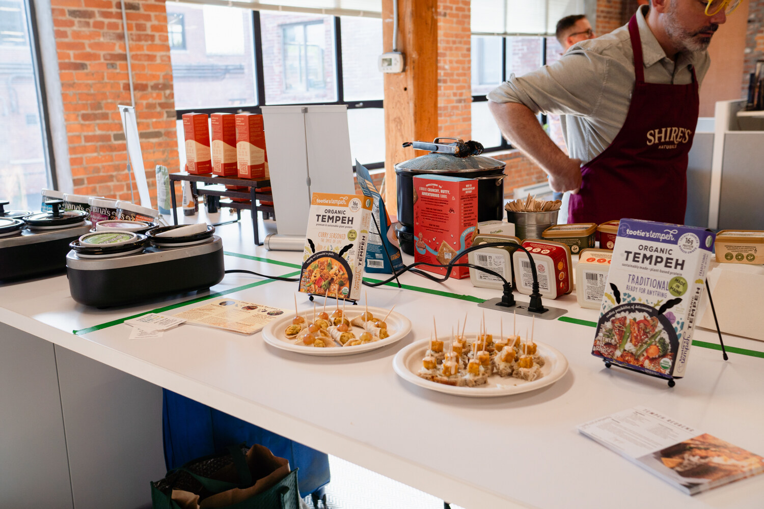 A sampling table with slow cookers, plates of bite-size samples and packages of organic tempeh, with a man in a maroon apron behind it.