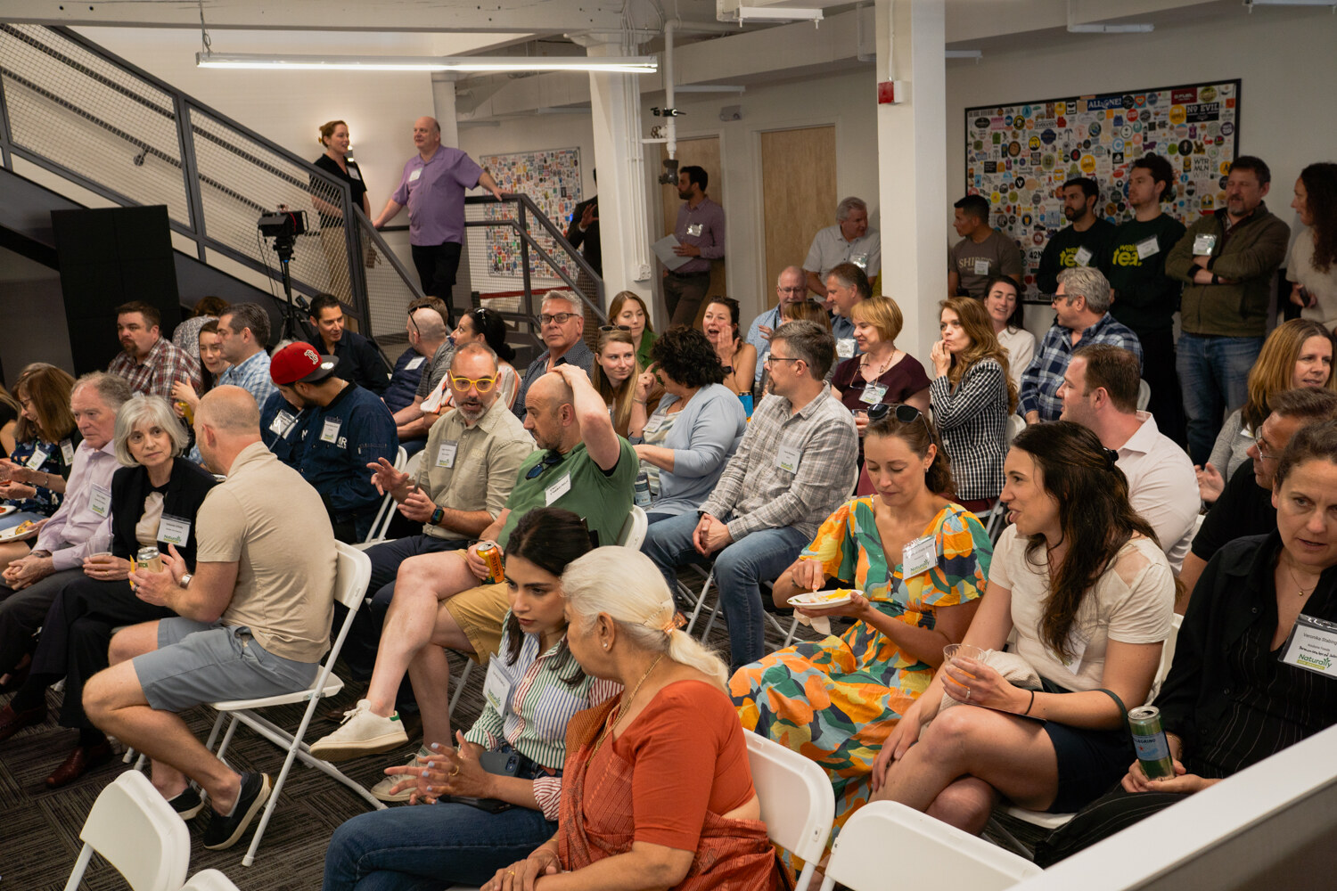 A packed audience seated in folding chairs, some holding plates of food, beneath a staircase.