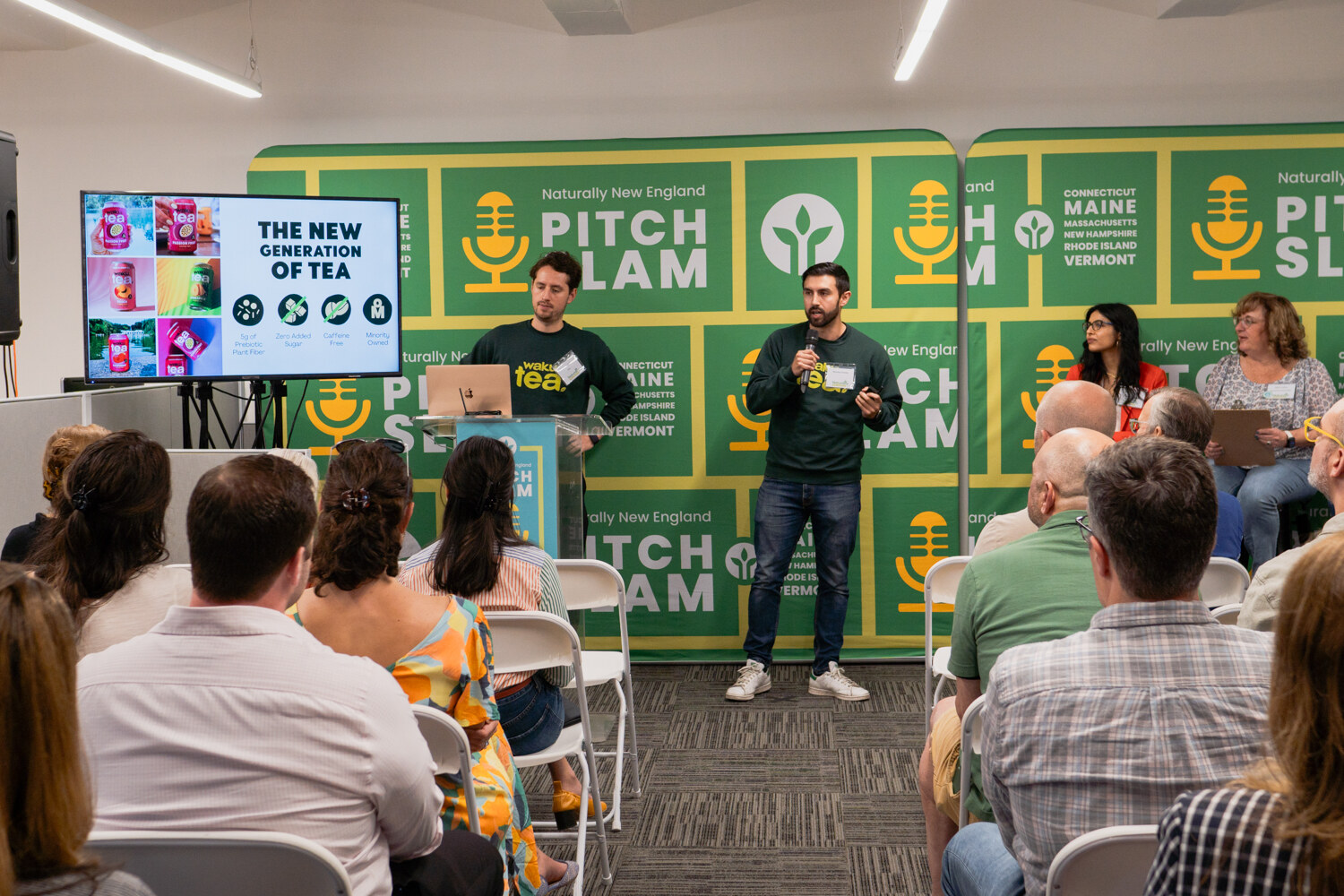 Two men in dark green sweatshirts present beside a screen reading The New Generation of Tea, with judges seated at right and an audience in the foreground.