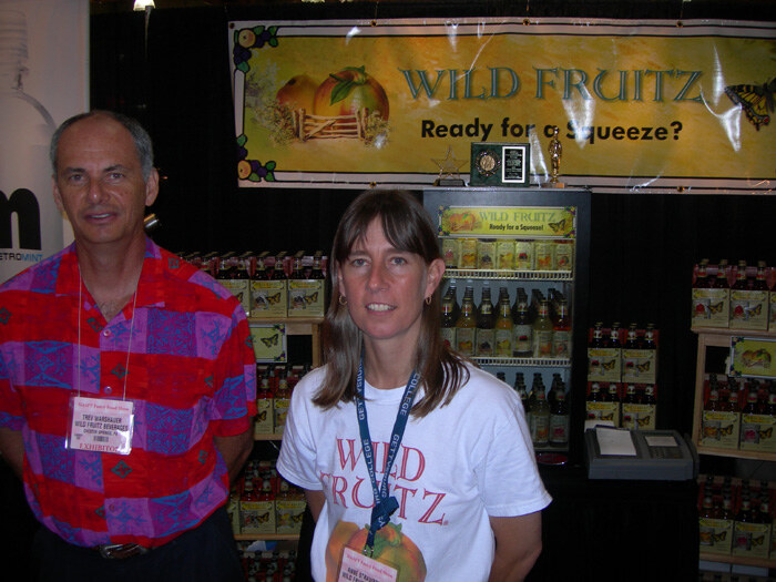 Trev Warshauer of Wild Fruitz Beverages, in a red patterned shirt, stands at left beside a woman in a white Wild Fruitz T-shirt in front of bottles and a Wild Fruitz banner.