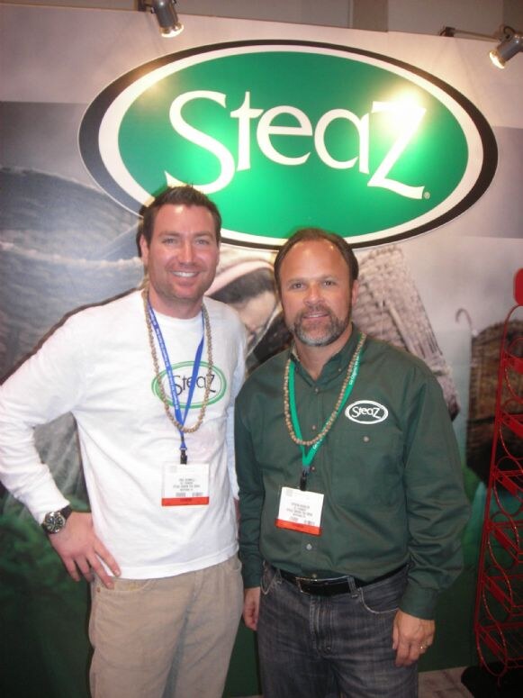 Two smiling men wearing beaded necklaces and show badges stand under a green oval Steaz sign. One wears a white Steaz shirt and the other a green Steaz button-up shirt.