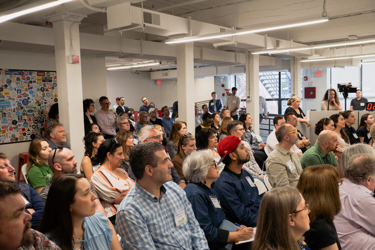 A wide view of a packed audience watching a presentation, with a sticker-covered wall and a camera tripod at the back.