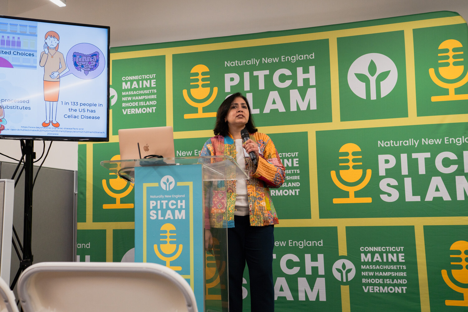 A woman in a colorful jacket speaks into a microphone at a clear podium in front of the green Pitch Slam backdrop and a screen with a cartoon illustration.