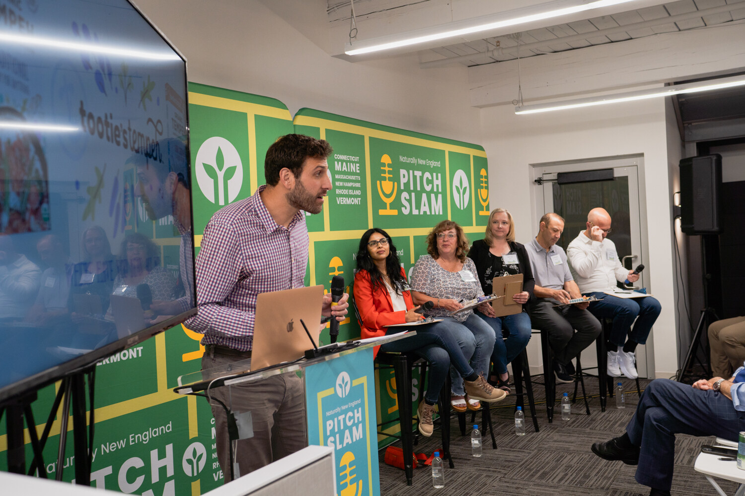 A man in a checked shirt stands next to a large screen showing the Tootie's logo while judges sit on stools.