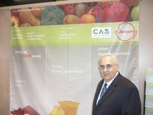A man in glasses, a dark suit and a striped tie stands in front of a Steinhauser Group banner that also shows the C.A.S. logo, fruit and vegetables, and lists purees, juices and powders.