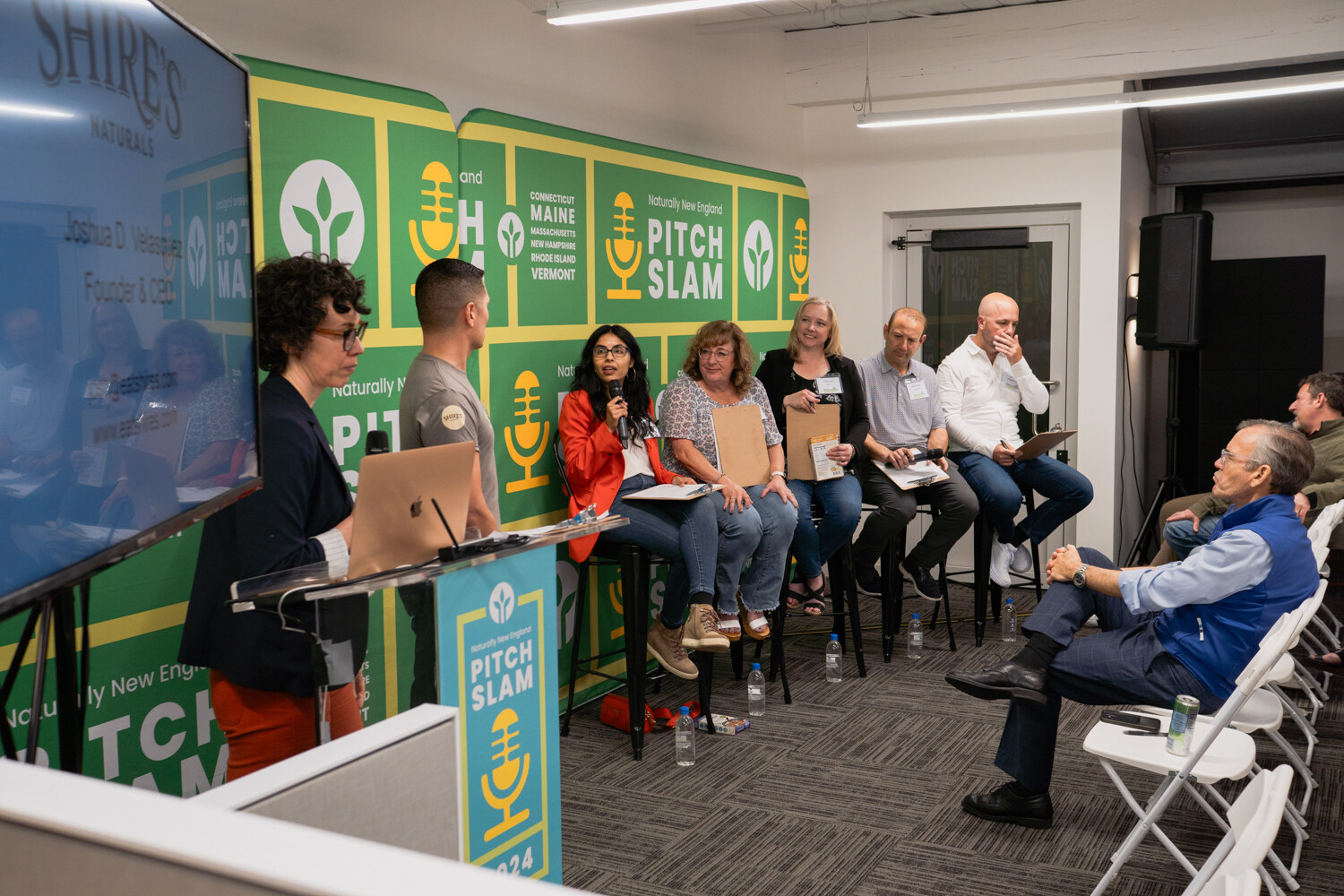 A man in a grey T-shirt and a woman in a navy blazer stand next to a screen with the Shire's Naturals logo while judges sit on stools.