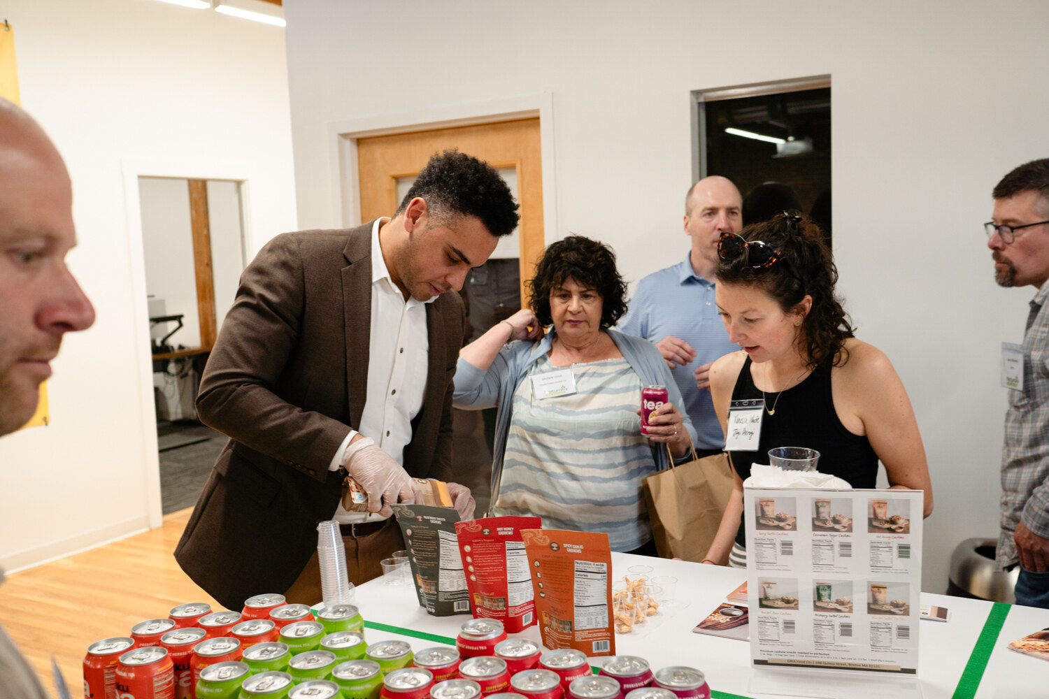 A man in a brown blazer and gloves reaches over a table of red and green cans while guests wait.