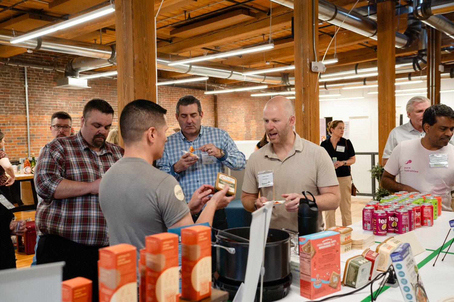 Several attendees stand at a table of orange boxes, pink cans and a slow cooker in a brick-walled room.
