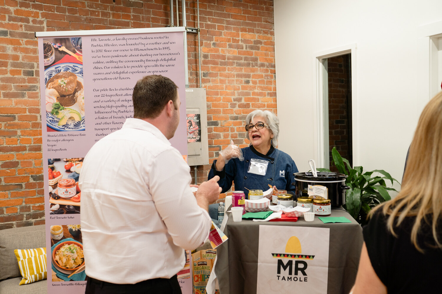 A man in a white shirt talks with a seated woman in a navy shirt behind a table with a Mr Tamole sign and slow cooker.