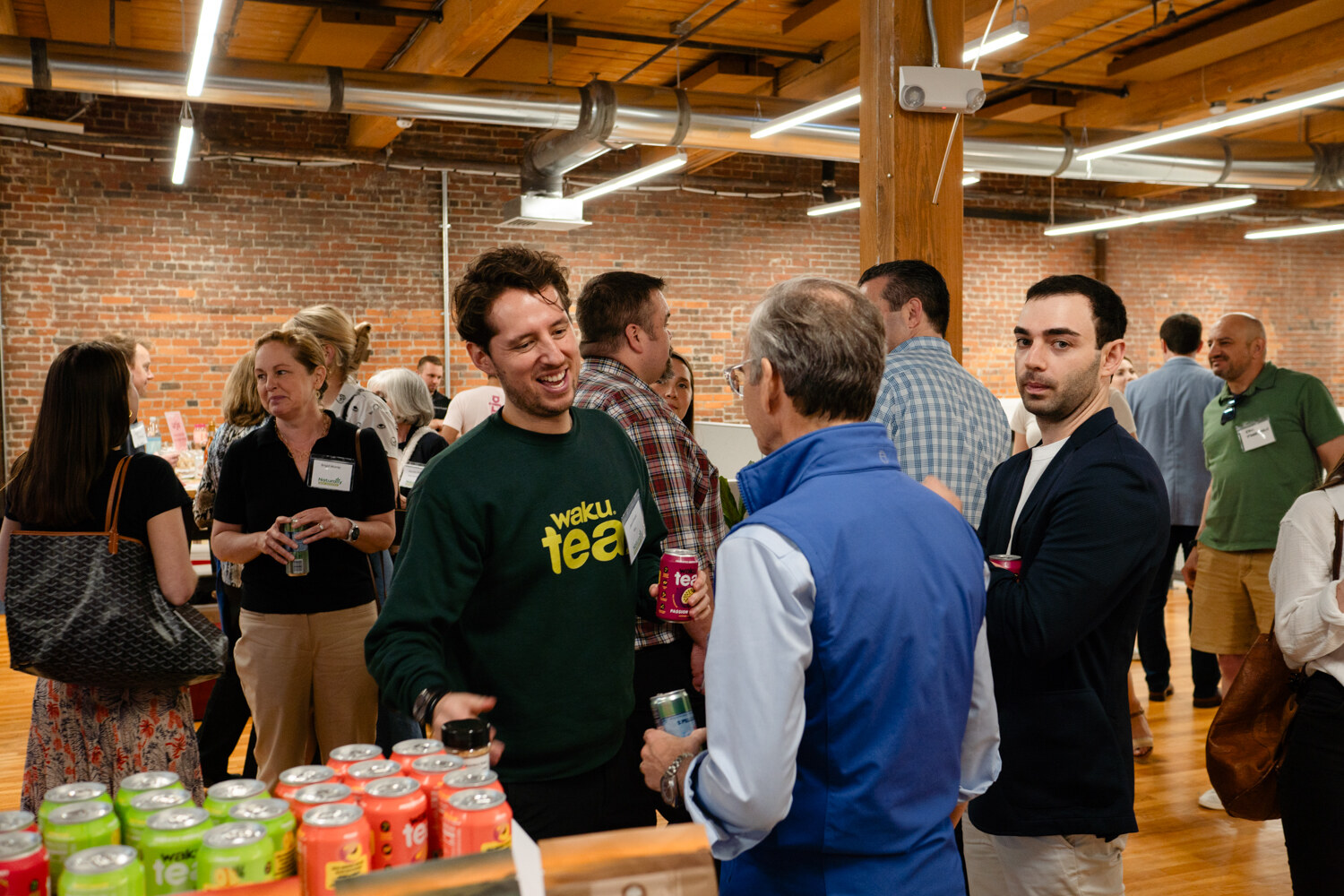 A man in a green Waku Tea sweatshirt laughs with an older man in a blue vest while other guests stand nearby and cans sit in the foreground.