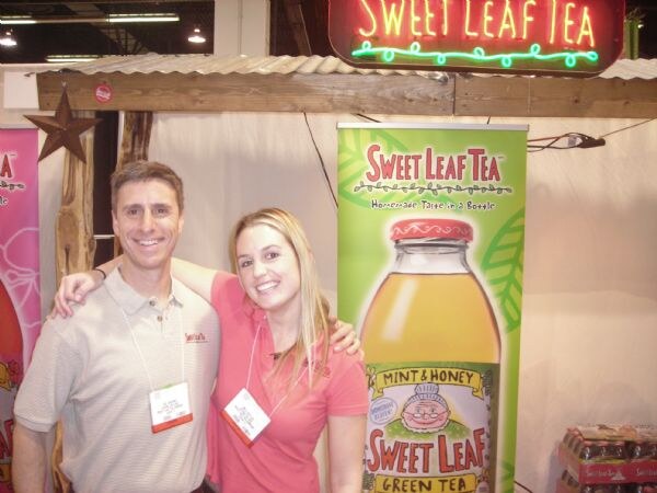 A smiling man in a gray polo and a woman in a pink shirt stand with their arms around each other. A Sweet Leaf Tea Mint & Honey Green Tea banner and a lit Sweet Leaf Tea sign are behind them.