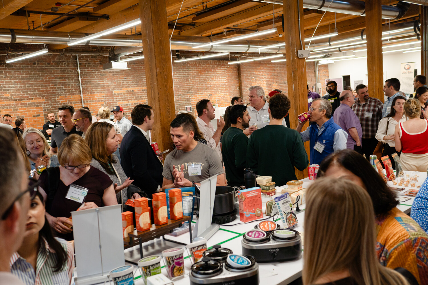 A crowd of attendees stands around tables of snacks and slow cookers in a room with brick walls and wooden beams.