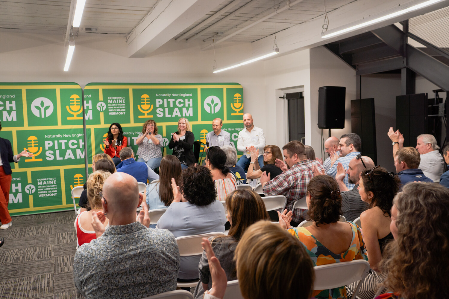 Judges sit on stools in front of the backdrop while the audience applauds in the foreground.