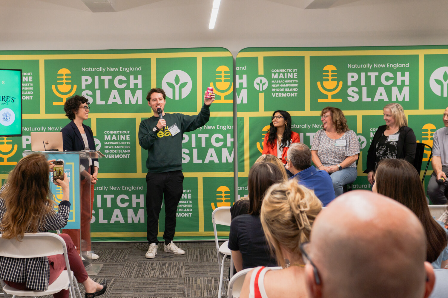 A man in a dark green sweatshirt holds a can above his head in front of the Pitch Slam backdrop while the host and judges look on.