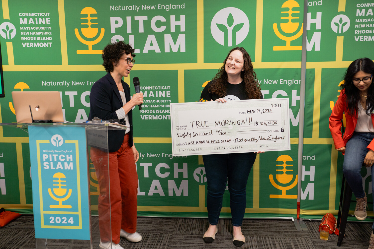 A woman in a navy blazer and a woman in a black T-shirt hold an oversized check made out to True Moringa in front of the Pitch Slam backdrop.