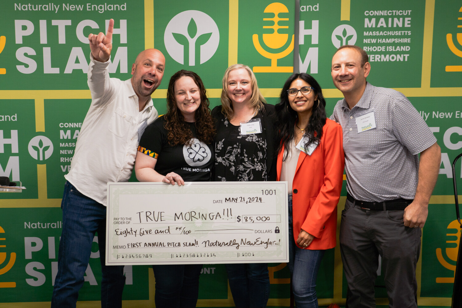 Five people stand behind an oversized True Moringa check, with a man on the left raising a hand in a rock gesture.